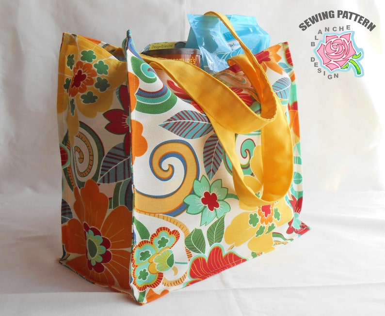 May include: A large, colorful tote bag with a floral pattern in shades of orange, yellow, red, green, and blue. The bag has yellow handles and is made of a sturdy fabric. The bag is sitting on a white surface.