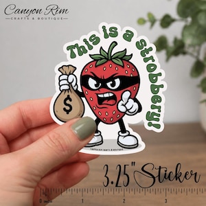 May include: A die-cut sticker featuring a cartoon strawberry with a black mask, holding a money bag. The text "This is a strobbery!" is in green. The sticker measures 3.25 inches.