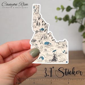 May include: A sticker in the shape of Idaho, featuring a detailed map with illustrations of landmarks and natural features. The sticker is approximately 3.1 inches in size, with a beige and blue color scheme. The text "Canyon Rim - Crafts & Boutique" is at the top.