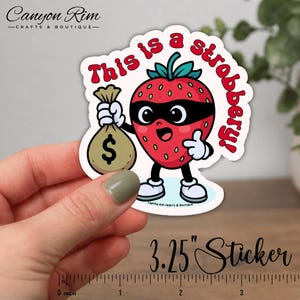 May include: A colorful sticker featuring a cartoon strawberry with a black eye mask, holding a money bag. The text "This is a strobbery!" is written above the strawberry. The sticker measures 3.25 inches.
