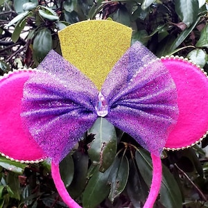May include: A pink headband with large, round, hot pink ears trimmed with gold beads. A large, glittery purple bow is centered, with a clear gem. A gold glitter crown sits above the bow, creating a whimsical accessory.