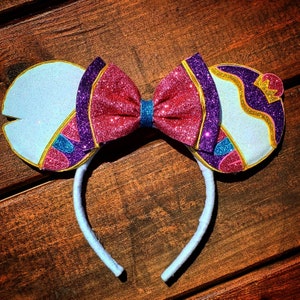 May include: A pair of white and purple glitter mouse ears with a pink and purple bow. The ears are decorated with gold and blue accents.