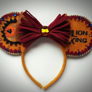 May include: Orange and brown felt mouse ears with a red bow and gold glitter. The ears have blue and gold embellishments and the right ear says "Lion King".