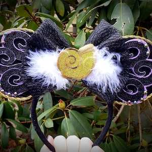 May include: Black headband with large, round ears. The ears are black with purple swirls, gold trim, and iridescent beads. A black bow and gold glitter seashell are centered between the ears, with white feathers.