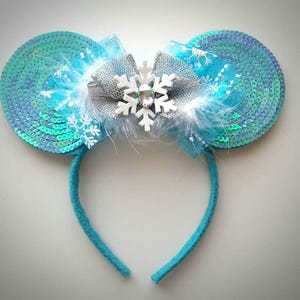 May include: Blue and silver sequined Minnie Mouse ears with a white snowflake bow and white feathers.
