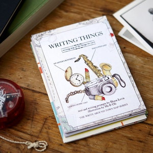 May include: A white book titled "Writing Things: A writing map of the things we carry" with illustrations of a saxophone, lipstick, a pocket watch, and a camera. The book is part of the "Write Around Town Map Series" and is by Shawn Levin and Me & Oli.
