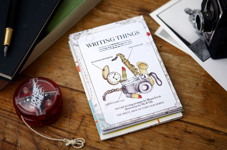 May include: A white book titled "Writing Things: A writing map of the things we carry" with illustrations of a saxophone, lipstick, a pocket watch, and a camera. The book is part of the "Write Around Town Map Series" and is by Shawn Levin and Me & Oli.