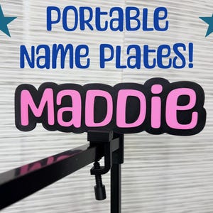 May include: A portable name plate with the name "MADDIE" in pink letters on a black background. The text "PORTABLE Name PLATES!" is in blue. Two blue stars are on the top corners. The name plate is attached to a black stand.