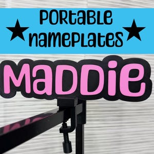 May include: A black nameplate with the name "MADDIE" in pink letters. The nameplate is mounted on a black stand. Above the nameplate is a blue banner with the words "PORTABLE namePLATES" and two black stars.