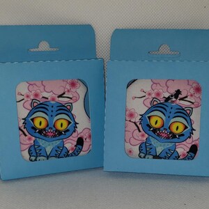 May include: Two pale blue gift boxes, each with a square window revealing a fabric square. The fabric displays a blue cartoon cat with yellow eyes, set against a pink floral backdrop. The boxes have a small hole at the top.