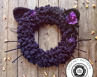 May include: A black crocheted cat wreath with a purple flower and spider accents. The wreath is on a wooden background with candy corn scattered around it. The text "Crochet Pattern The Hook Pusher digital download" is on a black circle.