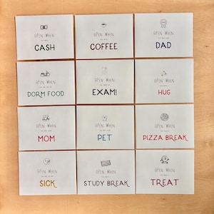 May include: Twelve white envelopes arranged in a grid, each featuring a small black icon, the text "OPEN WHEN," and a specific prompt in colored lettering: CASH, COFFEE, DAD, DORM FOOD, EXAM, HUG, MOM, PET, PIZZA BREAK, SICK, STUDY BREAK, and TREAT.