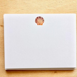 May include: A stack of white notecards with a seashell design in the upper center. The seashell is orange and brown. The notecards are on a light brown wooden surface.