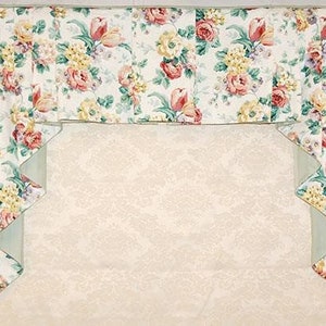 May include: A white valance with a floral pattern in shades of pink, green, and yellow. The valance has a scalloped edge and is trimmed with a green fabric.