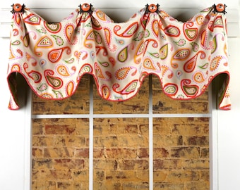 May include: A white valance with a paisley pattern in shades of orange, green, pink, and blue. The valance has a scalloped edge and is trimmed with a red and white ribbon. It is hung on a white window frame.