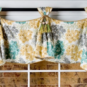 May include: A white valance with a floral pattern in shades of yellow, grey, and teal. The valance has a gold trim and wooden tassels with a light pink bead.