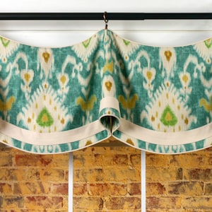 May include: A teal, white, and gold patterned valance with a white trim. The valance is hung on a black rod with gold finials. The valance is in front of a brick wall.