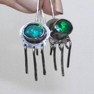 May include: A pair of silver dangle earrings with green gemstone accents. Each earring features a round green stone set in a silver bezel and five dangling silver strands.