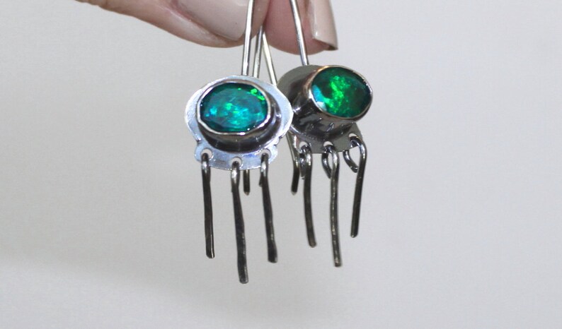 May include: A pair of silver dangle earrings with green gemstone accents. Each earring features a round green stone set in a silver bezel and five dangling silver strands.