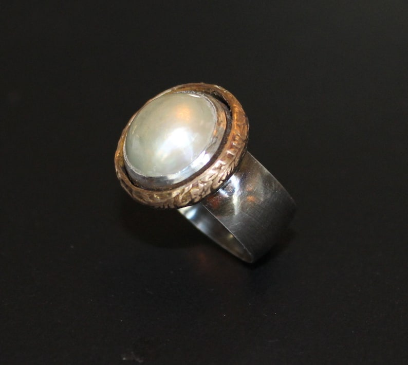 K&ouml;nnte beinhalten: Ein silberner Ring mit einem wei&szlig;en Perlmutt, das in eine goldene Fassung gefasst ist. Der Ring hat eine strukturierte Band.