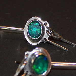 May include: A pair of silver earrings with a green gemstone set in a silver bezel. The earrings have a dangling design with a small silver chain.