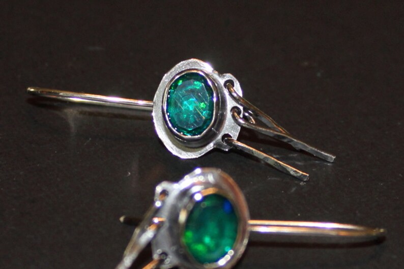 May include: A pair of silver earrings with a green gemstone set in a silver bezel. The earrings have a dangling design with a small silver chain.