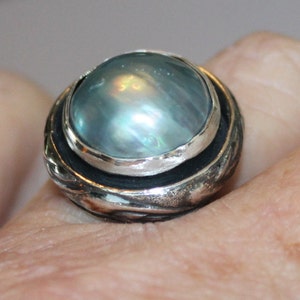 K&ouml;nnte beinhalten: Ein silberner Ring mit einem gro&szlig;en, runden, irisierenden blauen Edelstein, der in einer silbernen Fassung sitzt. Der Ring hat eine strukturierte Band mit einem silbernen und schwarzen Design.