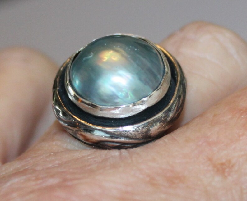 K&ouml;nnte beinhalten: Ein silberner Ring mit einem gro&szlig;en, runden, irisierenden blauen Edelstein, der in einer silbernen Fassung sitzt. Der Ring hat eine strukturierte Band mit einem silbernen und schwarzen Design.