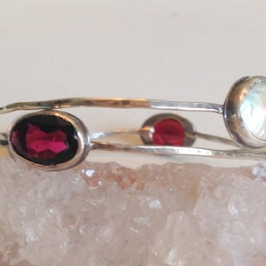 May include: A silver bangle bracelet featuring three stones: a large oval-shaped red garnet, a smaller round red garnet, and a round white pearl.