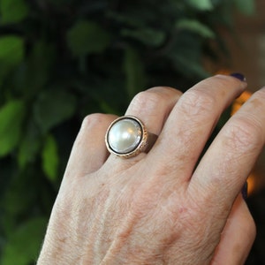 K&ouml;nnte beinhalten: Ein silberner Ring mit einem gro&szlig;en, wei&szlig;en Perlmutt, das in einer strukturierten, goldfarbenen Fassung sitzt. Der Ring wird an einem Finger getragen.