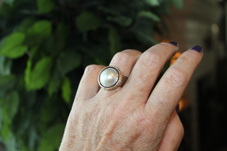 K&ouml;nnte beinhalten: Ein silberner Ring mit einem gro&szlig;en, wei&szlig;en Perlmutt, das in einer strukturierten, goldfarbenen Fassung sitzt. Der Ring wird an einem Finger getragen.