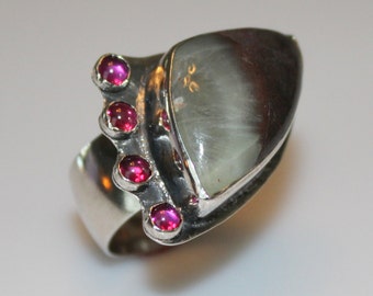 May include: A silver ring with a large, triangular, iridescent gemstone set in the center. The ring is accented with five small, pink gemstones.