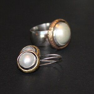 K&ouml;nnte beinhalten: Ein Set aus silbernen und goldenen Ohrringen und einem Ring mit Perlenakzent. Die Ohrringe sind ein Paar kleiner Creolen mit einer Perle in der Mitte. Der Ring ist ein schlichtes Band mit einer Perle, die in einer goldenen Fassung sitzt.