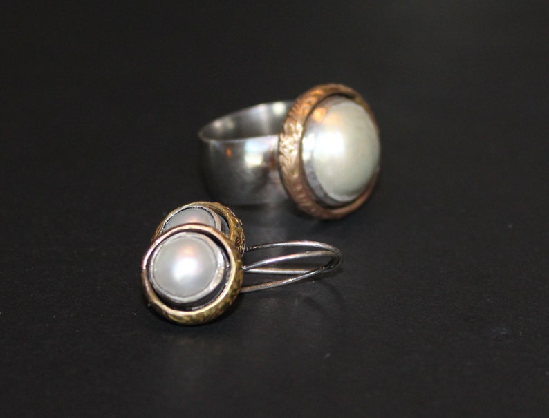 K&ouml;nnte beinhalten: Ein Set aus silbernen und goldenen Ohrringen und einem Ring mit Perlenakzent. Die Ohrringe sind ein Paar kleiner Creolen mit einer Perle in der Mitte. Der Ring ist ein schlichtes Band mit einer Perle, die in einer goldenen Fassung sitzt.