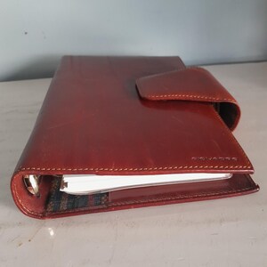 May include: A reddish-brown leather planner with a flap closure. The planner has a stitched border and a silver metal ring binder. The brand name "PIQUADRO" is visible on the front cover. The planner is closed, with the pages slightly visible.