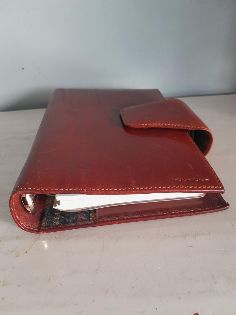 May include: A reddish-brown leather planner with a flap closure. The planner has a stitched border and a silver metal ring binder. The brand name "PIQUADRO" is visible on the front cover. The planner is closed, with the pages slightly visible.