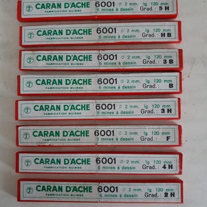 Op de afbeelding: Een verzameling Caran d'Ache 6001 potloodstiftendoosjes. Elke rode doos heeft witte tekst met informatie over de stift: diameter 2 mm, lengte 120 mm en hardheidsgraad. De dozen zijn gemarkeerd met "Fabrication Suisse".