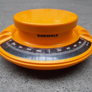 以下が含まれることがあります： オレンジ色のSOEHNLEキッチンスケール、円形デザイン。 最大1kgまで測定できる白黒の文字盤が付いています。 プラスチック製で、中央が盛り上がっています。