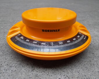 May include: An orange SOEHNLE kitchen scale with a circular design. The scale features a black and white dial with measurements up to 1 kg. The scale is made of plastic and has a raised center.