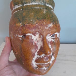Puede incluir: Una maceta de cerámica con forma de rostro, con base marrón, parte superior verde y detalles blancos. La maceta tiene un diseño de rostro sonriente con detalles. La maceta se sostiene en una mano.
