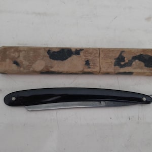 May include: A vintage straight razor with a black handle and a silver blade, and its original cardboard case. The case is rectangular with dark markings. The razor is curved and has a hook at the end.
