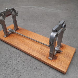 May include: A vintage metal and wood tool. The tool has two silver metal uprights with adjustable clamps, connected by a metal rod. The uprights are mounted on a rectangular wooden base. The base is a light brown color.
