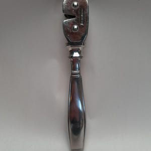 May include: A vintage, silver-toned can opener with a long, rounded handle and a distinctive cutting head. The head features a curved blade and two rivets. The handle has a decorative, ridged section near the top. The can opener is designed for kitchen use.