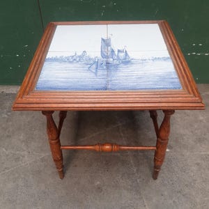 Op de afbeelding: Vierkante bijzettafel met een houten frame en poten. Het tafelblad heeft een blauw-wit tegelontwerp met een zeegezicht met zeilboten. De tafel is ongeveer 61 cm vierkant en heeft een warme bruine afwerking.
