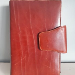 May include: A reddish-brown leather journal with a flap closure. The journal has visible grain and stitching details. The cover is a solid color with a rectangular shape. The journal is a classic style.