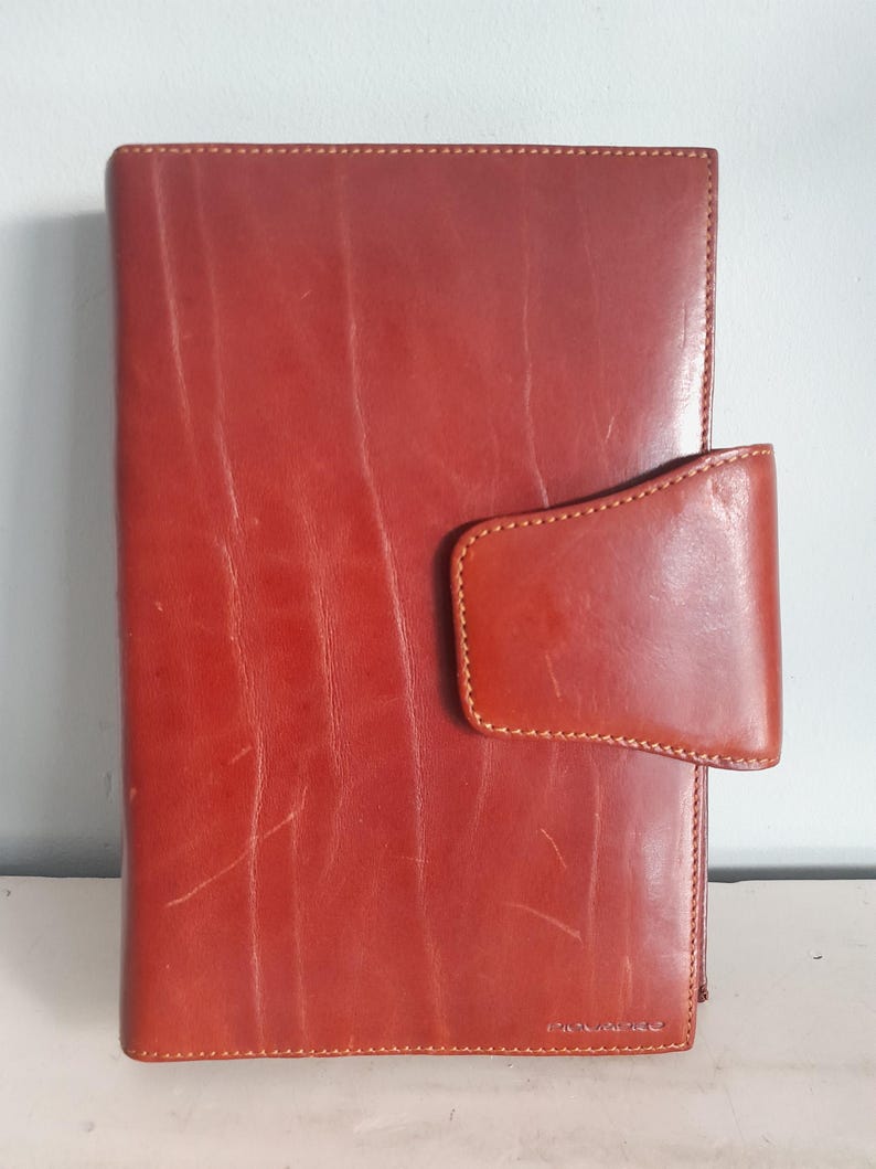 May include: A reddish-brown leather journal with a flap closure. The journal has visible grain and stitching details. The cover is a solid color with a rectangular shape. The journal is a classic style.