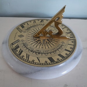 May include: A brass sundial with a compass and a calendar on a white marble base. The sundial has Roman numerals and a compass rose. The calendar shows the months of the year and the days of the week.