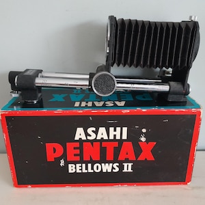 Puede incluir: Un accesorio de cámara vintage Asahi Pentax Bellows II. El fuelle negro está montado en un sistema de rieles metálicos con marcas de medición. El artículo descansa sobre su caja original, que tiene detalles en rojo, negro y verde azulado, con el nombre de la marca en un lugar destacado.
