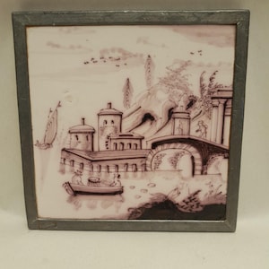 Könnte beinhalten: Quadratische Keramikfliese mit einer sepiafarbenen Landschaft in einem Metallrahmen. Das Bild zeigt eine Uferlandschaft mit Gebäuden, einer Brücke und einem Boot. Der Himmel hat ein paar Vögel. Die Fliese ist wahrscheinlich dekorativ.