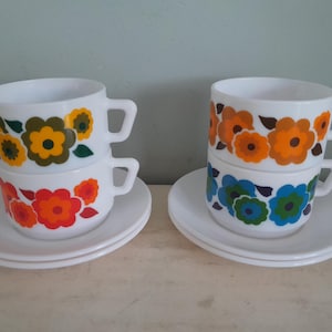 May include: Two sets of vintage white teacups and saucers. Each cup is decorated with a floral pattern in orange, yellow, green, and blue. The cups are stacked on top of each other, with a saucer beneath each set. The background is a light green.
